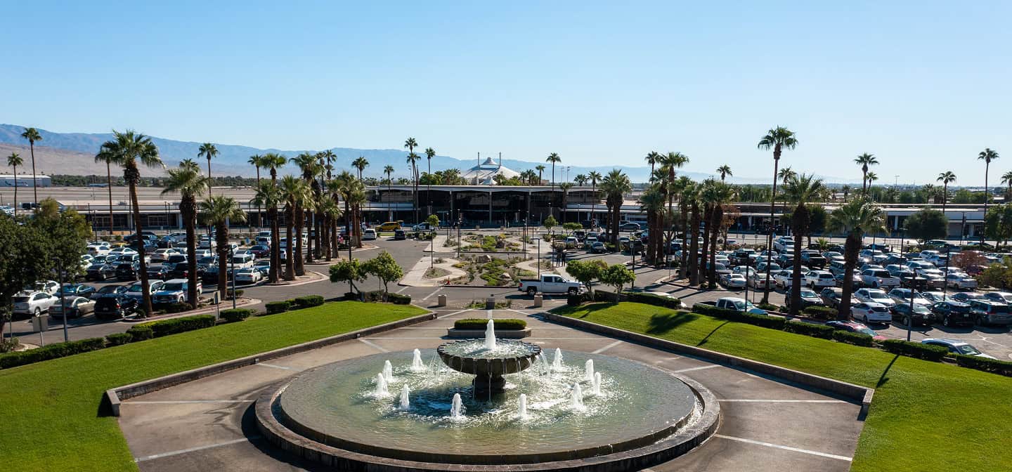Elevated view of an airport entrance with a large circular fountain in the foreground, surrounded by green grass. Beyond are parking lots filled with cars, numerous tall palm trees, and the airport terminal building with a white tent-like roof. Distant mountains are visible under a clear blue sky.